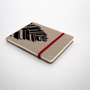 Libreta forrada en lino tono moca con estampado negro, elástico de cierre rojo, cosida y encuadernada a mano sobre fonde grisáceo de la marca Leaftones creada por Diana Herrera Design